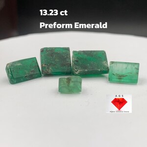 May include: Six green emerald gemstones, preformed and unpolished, weighing a total of 13.23 carats. The gemstones are arranged on a white surface. The text "AGS Afghan Gemstone Source" is visible in the lower right corner of the image.