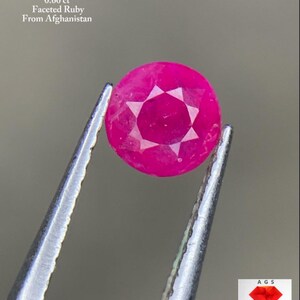 Puede incluir: Un rubí facetado, sostenido por unas pinzas, con el texto "0.60 ct Faceted Ruby From Afghanistan". El rubí es de color rosa vibrante con un patrón en forma de estrella en el centro. La imagen incluye el logotipo de "AGS Afghan Gemstone Source".
