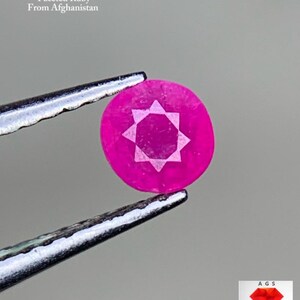 Puede incluir: Un rubí facetado de corte redondo, sostenido por unas pinzas. El rubí es de color rosa vibrante con un diseño en forma de estrella en el centro. El texto de la imagen dice "0.22 ct Faceted Ruby From Afghanistan."