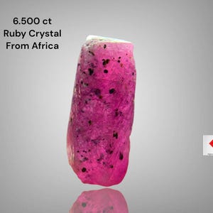 May include: A vibrant pink ruby crystal from Africa, weighing 6.500 ct. The gemstone has a rough, natural texture with dark inclusions. The text "6.500 ct Ruby Crystal From Africa" is visible.