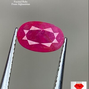 Puede incluir: Un rubí facetado de Afganistán, sujeto con pinzas. La piedra roja oscura de forma ovalada tiene un patrón de facetas en forma de estrella. El texto de la imagen dice "1,01 ct Rubí Facetado De Afganistán."