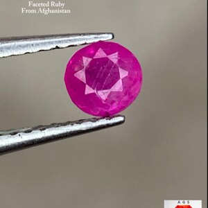 May include: A faceted, round-cut ruby, held by tweezers. The gemstone is a vibrant pink-red color. Text in the image reads: "0.24 ct Faceted Ruby From Afghanistan."