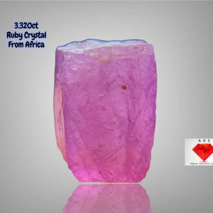 Puede incluir: Cristal de rubí rosa vibrante de África, con un peso de 3,320 ct. La gema tiene forma rectangular con una textura ligeramente rugosa y una superficie reflectante. La imagen incluye el texto "3.320ct Ruby Crystal From Africa" y un logotipo.