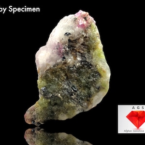 May include: A rough, pink ruby specimen with green and black inclusions. The specimen is on a black background. The text "Ruby Specimen" is at the top of the image. The text "AGS Afghan Gemstone Source" is at the bottom of the image.