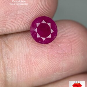 Puede incluir: Una gema de rubí facetada y redonda, de color rojo intenso. La gema pesa 1,87 quilates y es de Afganistán. La imagen incluye el texto "1.87 ct Faceted Ruby From Afghanistan."