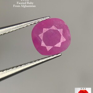 Puede incluir: Un rubí facetado, talla cojín, sujeto con pinzas. La gema es de un color rojo-rosa vibrante, con un patrón en forma de estrella tallado en su superficie. El texto dice "1.71 ct Faceted Ruby From Afghanistan."