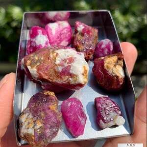 Pode incluir: Uma coleção de rubis brutos de 420 ct do Afeganistão em uma bandeja de metal retangular. As pedras variam em tamanho e forma, apresentando texturas cristalinas vermelho-escuras, rosas e brancas com superfícies naturais, não polidas e irregulares.