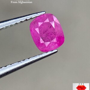Może przedstawiać: Fasetowany, żywy różowy rubin o masie 0,44 ct jest trzymany przez pęsetę. Kamień szlachetny, pochodzący z Afganistanu, prezentuje symetryczny szlif z widocznymi fasetami. Obraz zawiera tekst wskazujący pochodzenie i wagę rubinu.
