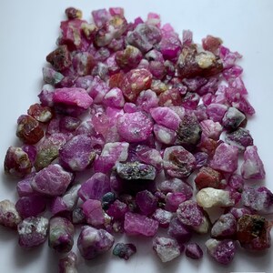 Pode incluir: Uma coleção de pedras preciosas de rubi bruto em tons de rosa, vermelho e branco. A imagem inclui o texto "30.2 ct Rough Ruby Lot". As pedras preciosas estão espalhadas sobre uma superfície branca.