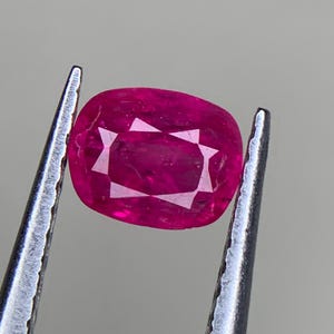 Pode incluir: Um rubi de corte almofada, preso por pinças. O rubi vermelho profundo exibe facetas internas que refletem a luz. A pedra preciosa é o ponto focal, destacando sua clareza e cor.
