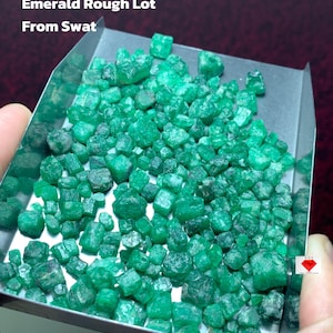 May include: A metal tray overflowing with raw emeralds. The gemstones are a vivid green hue and of varying sizes. Text on the image reads: "325.45 ct Emerald Rough Lot From Swat."