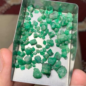 May include: A collection of rough emeralds, with a total weight of 38.65 carats, displayed in a small metal tray. The emeralds are a vibrant green color and appear to be uncut. Text on the image reads "Emerald Rough Lot From Swat".