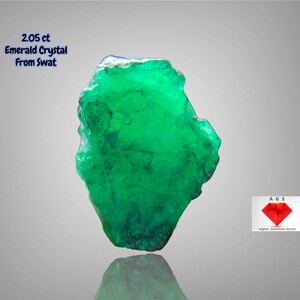 May include: A vivid green emerald crystal from Swat, weighing 2.05 ct. The rough gemstone has a natural, irregular shape and a reflective surface. The image also includes the logo of Afghan Gemstone Source.