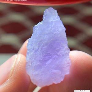 Puede incluir: Un cristal de hackmanita de color púrpura claro de Afganistán, que pesa 23,82 ct. La gema en bruto tiene una superficie texturizada y se sostiene entre los dedos. La imagen incluye el texto "23.82 ct Hackmanite Crystal From Afghanistan".