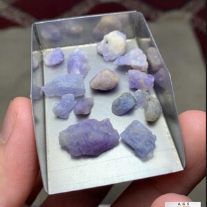 May include: A collection of 40 ct rough Hackmanite stones from Afghanistan displayed in a metal scoop. The raw, unpolished minerals exhibit varying shades of light purple, lavender, and off-white, with irregular, jagged shapes and textures.