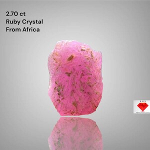 Puede incluir: Cristal de rubí rosa en bruto de África, con un peso de 2,70 ct. La gema tiene una forma irregular con una superficie reflectante. La imagen incluye el texto "2.70 ct Ruby Crystal From Africa".