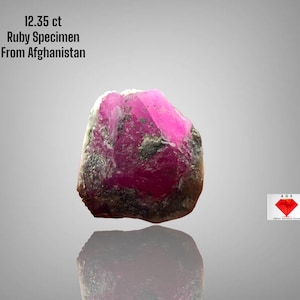 May include: A raw ruby specimen from Afghanistan, weighing 12.35 ct. The gemstone has a vibrant pink hue with darker, mottled areas. The image includes text indicating the origin and weight of the ruby.