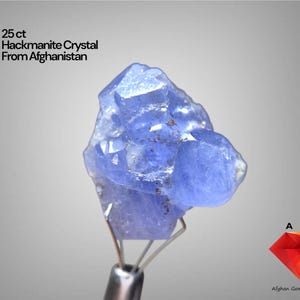 Puede incluir: Cristal de hackmanita de 25 ct. de Afganistán. El cristal es de un tono azul vibrante, con una textura rugosa y natural. La imagen incluye el texto "25 ct Hackmanite Crystal From Afghanistan."