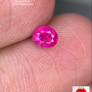 以下が含まれることがあります： 鮮やかなピンク色の、約0.40ctのファセット加工されたオーバルカットのルビー。アフガニスタン産であることを示すテキストとともに、肌色の背景に宝石が展示されています。