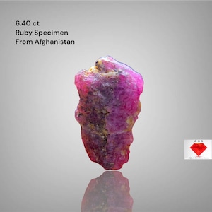 Peut inclure: Spécimen de rubis brut d'Afghanistan, pesant 6,40 carats. La pierre précieuse est d'une couleur rouge-rosé vibrante avec une surface rugueuse et texturée. Le texte indique l'origine et le poids du rubis.