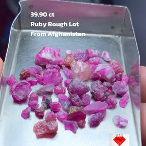 Puede incluir: Un conjunto de gemas de rubí en bruto en una bandeja plateada. Las gemas son de varios tonos de rosa y rojo, con algunas inclusiones blancas. El texto en la bandeja dice "39.90 ct Ruby Rough Lot From Afghanistan."