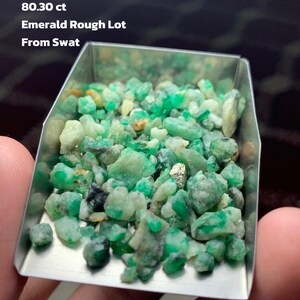 Puede incluir: Un conjunto de esmeraldas en bruto de color verde, presentadas en una pequeña bandeja metálica. El texto en la imagen dice "80.30 ct Emerald Rough Lot From Swat".