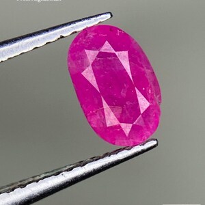 Puede incluir: Un rubí facetado de forma ovalada, de color rosa vibrante, sostenido por unas pinzas. La gema, con un peso de 0,70 ct, muestra facetas internas y está etiquetada como "From Afghanistan". La imagen incluye el logotipo "AGS".