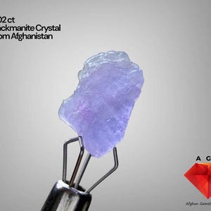 Puede incluir: Un cristal de Hackmanita de color púrpura claro de Afganistán, sostenido por una herramienta de metal. El cristal es de 3,02 ct. La imagen también incluye el texto "3.02 ct Hackmanite Crystal From Afghanistan" y un logotipo de diamante rojo con el texto "Afghan Gemstone Source."
