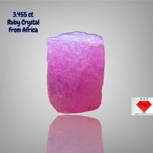 Puede incluir: Cristal de rubí rosa vibrante de África, 3,455 ct, sobre un fondo gris claro. La gema tiene forma rectangular con superficie texturizada y base reflectante. El texto indica origen y peso del cristal. También incluye un logotipo.