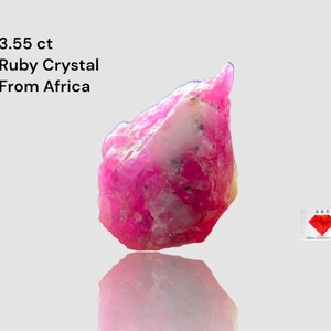 Puede incluir: Cristal de rubí rosa vibrante de África, con un peso de 3,55 ct. La gema en bruto tiene una superficie texturizada con inclusiones blancas. También incluye el texto "Ruby Crystal From Africa".