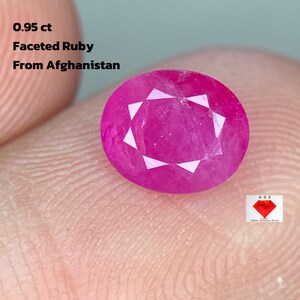 Puede incluir: Una gema de rubí facetada de corte redondo, de aproximadamente 0,95 quilates, en un tono rosa vibrante. La gema se sostiene entre los dedos, con texto que indica su origen afgano. Se ve un pequeño logotipo.