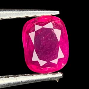 May include: A vibrant, oval-shaped ruby gemstone, held by tweezers against a black background. The ruby displays a deep pink hue with a star-like white pattern in the center, showcasing its facets and clarity.