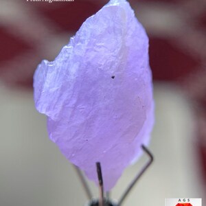 Puede incluir: Primer plano de un cristal de Hackmanita de 16,15 ct de Afganistán. El cristal es de color lavanda pálido y tiene una forma rugosa e irregular. El texto "16.15 ct Hackmanite Crystal From Afghanistan" es visible.