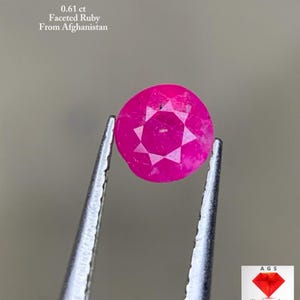 Könnte beinhalten: Ein facettierter, runder Rubin, gehalten von einer Pinzette. Der Rubin hat eine leuchtende rosa-rote Farbe mit einem Sternmuster in der Mitte. Der Text auf dem Bild lautet "0,61 ct Facettierter Rubin aus Afghanistan."