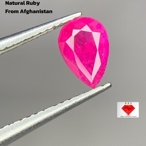 Op de afbeelding: Een gefacetteerde, peervormige natuurlijke robijn, vastgehouden door een pincet. De felroze edelsteen is gelabeld met "0.600 ct Natural Ruby From Afghanistan". De afbeelding bevat ook een logo voor "Afghan Gemstone Source".
