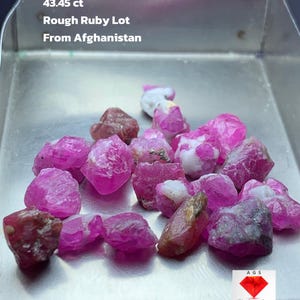 Puede incluir: Un lote de rubíes en bruto, con un peso total de 43,45 ct. Las piedras son de un color rosa vibrante, con algunas inclusiones blancas y marrones. El texto de la imagen dice "Rough Ruby Lot From Afghanistan."