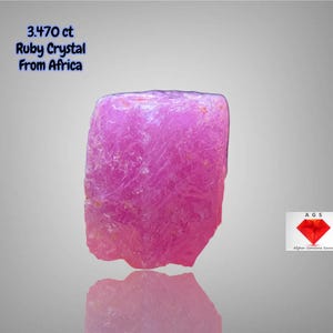 Puede incluir: Cristal de rubí rosa vibrante de África, con un peso de 3,470 ct. La gema en bruto tiene una superficie texturizada y se refleja en la superficie inferior. También incluye el texto "3.470 ct Ruby Crystal From Africa".