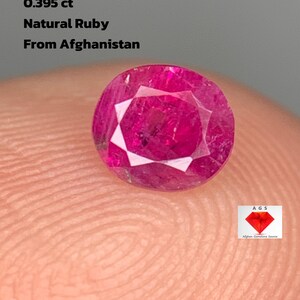 May include: A faceted, round natural ruby gemstone from Afghanistan, weighing 0.395 ct. The ruby is a deep pinkish-red colour with a brilliant cut, displayed on a fingertip. Text on the image reads: "0.395 ct Natural Ruby From Afghanistan."