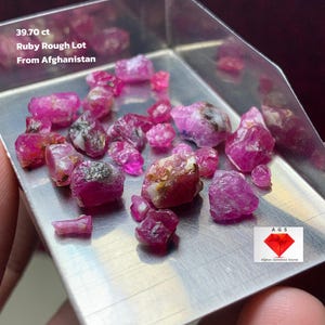 Puede incluir: Un lote de piedras preciosas de rubí en bruto, con un peso de 39,70 ct, presentado en una pequeña bandeja de metal. Los rubíes son de un color rojo-rosado vibrante, con algunas inclusiones naturales. El texto en la imagen dice "Ruby Rough Lot From Afghanistan."