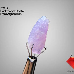 Puede incluir: Cristal de hackmanita de color púrpura claro de Afganistán, de 5,74 ct, sostenido por un instrumento metálico. El cristal tiene una superficie rugosa y texturizada sobre un fondo gris. La imagen también incluye el logotipo de Afghan Gemstone Source.
