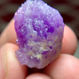 Puede incluir: Cristal de hackmanita morado en bruto de Afganistán, de 24,47 ct. La piedra tiene una superficie texturizada con zonas de color más claro. La imagen incluye el texto "24.47 ct Hackmanite Crystal From Afghanistan."
