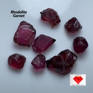 Op de afbeelding: Een verzameling van acht ruwe, onbewerkte rhodoliet granaat edelstenen. De granaten zijn in verschillende tinten roze en rood. De granaten liggen op een wit oppervlak. De tekst "Rhodolite Garnet" staat in de linkerbovenhoek van de afbeelding. De tekst "AGS Afghan Gemstone Source" staat in de rechterbenedenhoek van de afbeelding.