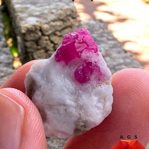 May include: A 33 ct ruby specimen featuring a vibrant, translucent deep pink crystal embedded within a rough, opaque white and grey mineral matrix. The text 33 ct Ruby Specimen is visible at the top, with an A G S logo at the bottom right.