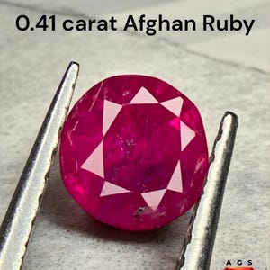 Op de afbeelding: Een dieproze-rode, rond geslepen Afghaanse robijn, vastgehouden door een pincet. De robijn heeft witte facetten. De tekst bovenaan luidt "0,41 karaat Afghaanse Robijn". Het logo "AGS Afghan Gemstone Source" staat rechtsonder.