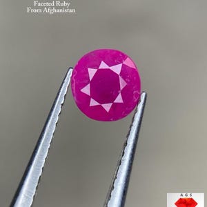 Puede incluir: Una gema de rubí redonda y facetada, sujeta con pinzas. La gema es de un color rojo-rosa vibrante con un patrón de facetas en forma de estrella. El texto en la parte superior dice "0,90 ct Rubí Facetado De Afganistán."