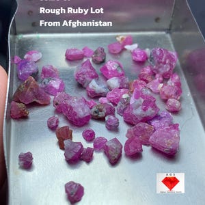 Puede incluir: Un conjunto de gemas de rubí en bruto en tonos rosados y rojizos, presentadas en una bandeja metálica. El texto "33.20 ct Rough Ruby Lot From Afghanistan" es visible. Las gemas sin cortar tienen un aspecto natural.