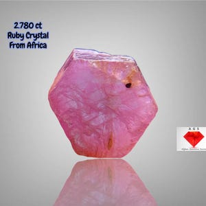 Puede incluir: Cristal de rubí rosa de África, con un peso de 2,780 ct. La gema tiene una forma facetada y una superficie reflectante. La imagen incluye el texto "2.780 ct Ruby Crystal From Africa" sobre un fondo gris degradado.