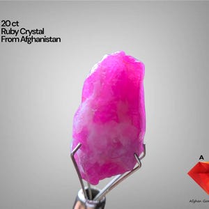 Puede incluir: Un vibrante cristal de rubí rosa de Afganistán, con un peso de 20 ct. La gema está sujeta por un soporte de metal plateado, mostrando su forma natural e irregular. El texto "20 ct Ruby Crystal From Afghanistan" está presente.