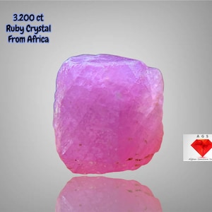 Könnte beinhalten: Ein leuchtend rosa Rubinkristall aus Afrika, mit einem Gewicht von 3,200 ct. Der Edelstein hat eine raue, natürliche Textur und wird vor einem grauen Farbverlaufshintergrund präsentiert. Der Text lautet "3.200 ct Ruby Crystal From Africa."