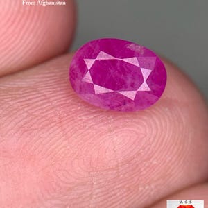Puede incluir: Se muestra una gema de rubí facetada, de forma ovalada y de un vibrante tono magenta. La gema es de 1,86 ct y procede de Afganistán. El texto "1.86 ct Faceted Ruby From Afghanistan" es visible.